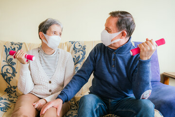 Adult couple with facemasks sitting on a sofa and working out with small dumbbells