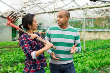 Naklejka premium Farmers man and woman with rake smiling and talking to each other at greenhouse farm