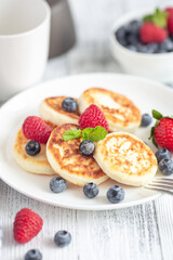 Cottage cheese pancakes with fresh berries, cup of coffee and coffee maker on white wooden table. Tasty breakfast food. Vertical photo - Image