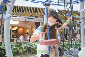 Cute little Asian boy child wearing washable face mask climb on jungle gym at pulbic indoor playground with his father during covid-19 outbreak, New normal lifestyle concept