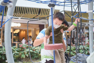 Cute little Asian boy child wearing washable face mask climb on jungle gym at pulbic indoor playground with his father during covid-19 outbreak, New normal lifestyle concept - Powered by Adobe