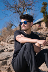 boy model sitting on rocks wearing black t-shirt black pant, sunglasses and looking away