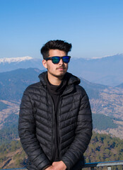 Indian ethnicity boy wearing black jacket and blue sunglasses looking at camera standing on top of mountain