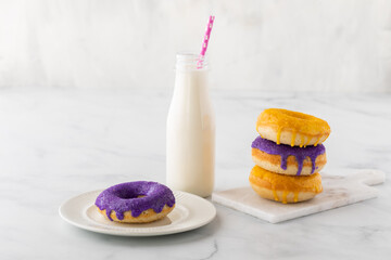 Close up of a stack of homemade donuts, a bottle of milk with a straw and a purple donut on a plate in front.