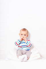 Funny caucasian baby boy in striped t-shirt sitting on pillow mountain with cookie in hand. White brick wall background with copy space. 