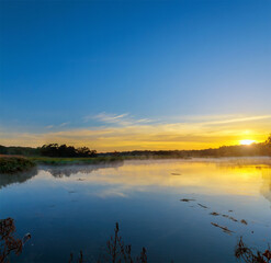 Serene lake at sunrise in forested, sun reflecting in the water with fog