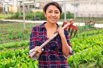 Attractive woman gardener worker standing with rake and smiling at greenhouse farm