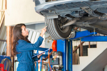 Asian woman mechanic holding a clipboard of service order and  looking at the tire to checking and assembles the wheel. Mechanic fixing car suspension with car lifting.