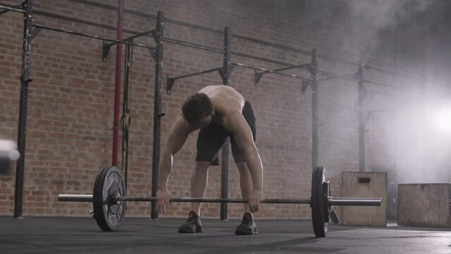 Wide shot of young determinate Caucasian sportsman wearing sport clothing and sneakers coating hands with powder and starting lifting up heavy barbell in gymWide shot of young determinate Caucasian sp