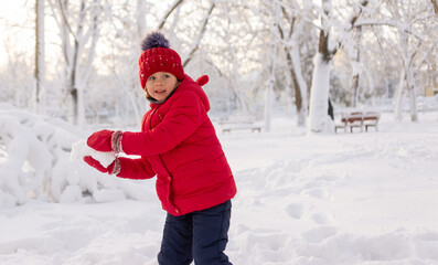 A little girl in mittens, a red jacket and a knitted hat throws snowballs in the park in winter....