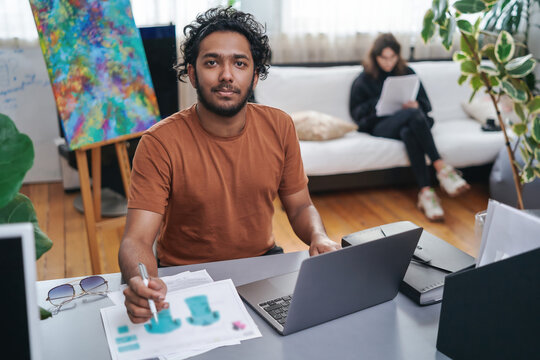 Cheerful male designer with curly hairs in casual attire sits at table and designing concept of shirts using a laptop and a pen.