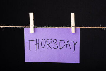 Thursday word written on a Purple color sticky note hanging with a wire on black background.