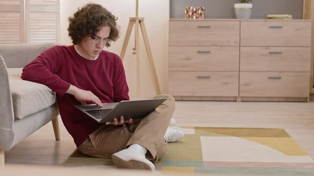 Full shot of curly dark-haired Caucasian guy wearing glasses sitting on floor, leaning back on sofa, using laptop computer, Pembroke Welsh Corgi dog chasing toy ball running pass him