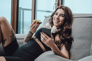 Portrait of a happy sportswoman with earphone and telephone sits on sofa holding sandwich and...
