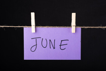 June word written on a Purple color sticky note hanging with a wire on black background.