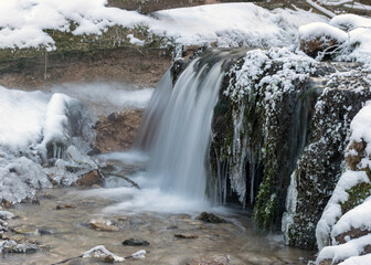 landscape with a small waterfall and flooded water, frozen icicles, stones covered with green moss, ice and snow