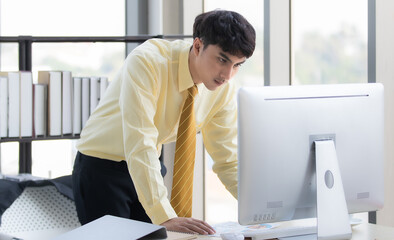 Young business man Asians, wearing a yellow long-sleeved shirt and tie, stand up, thoughtfully staring at the computer in the white room