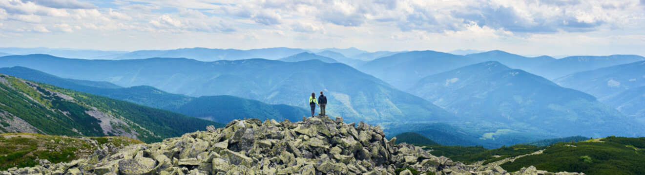 Panorama of Carpathian mountains with green forests, rear view pair is holding hands and enjoying powerful view. Man and woman is standing on top of cliff under the sky with cumulus clouds