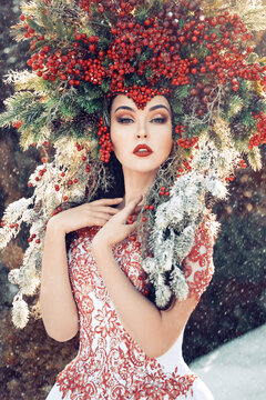 Fantasy Winter Queen Standing In The Forest In Falling Snow, A Beautiful Woman In The Crown Of Red Berries And Fur Branches