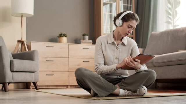 Full shot of young Caucasian woman wearing over-ear wireless headphones, sitting with legs crossed on floor in living room, using tablet computer, smiling, looking pleased
