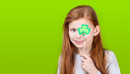 Portrait of little smiling caucasian red-haired girl covering her eye with shamrock clover leaf on...
