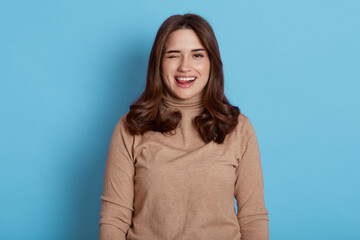 Fototapeta premium Young beautiful woman looking at camera, wearing casual clothes, dark haired positive female smiling, funny girl posing near blue wall, shows tongue and winks,