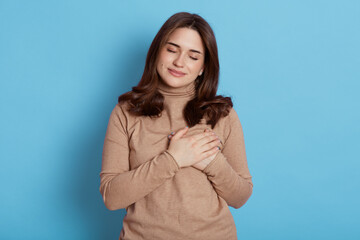 European emotional calm woman with closed eyes, dark haired lady keeps hands on chest, girl expresses her sympathy to someone, being thankful to somebody, isolated over beige background.