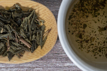 Cuchara de madera con hojas secas de té verde y taza con el poso del té sobre una mesa de madera.