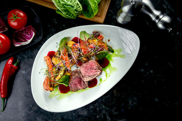 Medium rare beef medallions with vegetable garnish, served on a white plate. Dark marble background. Barbecue meat, restaurant food. Tasty steak