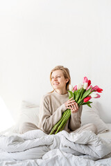 Happy woman sitting on the bed wearing pajamas holding tulip flowers bouquet