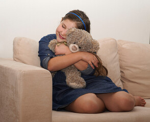 Little girl sitting on the sofa near wall in empty room and holding Teddy bear. Autism and loneliness concept.