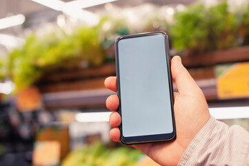 Mock-up of a smartphone with a white screen in the hands of a man. Phone on the background of in a supermarket. Environmentally friendly and healthy food.
