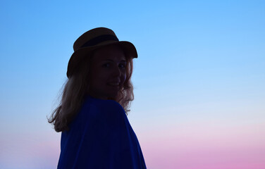 Young woman wearing straw hat and beach wrap against the sky at sunset in pink and blue pastel colors. Woman is looking over her shoulder at camera. Copy space. Silhouette