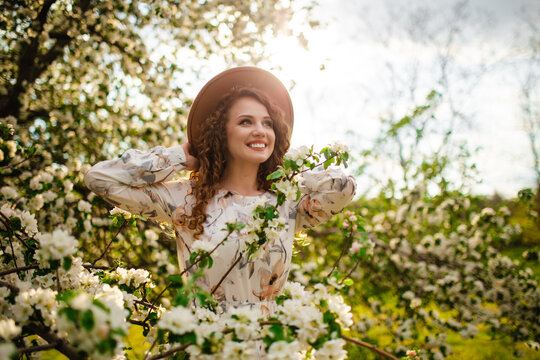 Portrait of a young girl with dark hair smiling and having fun in the middle of a blooming tree surrounded by lots of white flowers. Female in beige hat and white dress at the spring park.