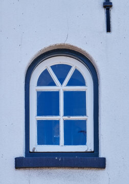 An old wooden window of a lighthouse. The green and white paint is peeling off. A classic semi-circular arched window in a white wall with iron lightning conductors