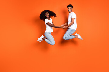 Full size photo of young good mood smiling afro couple on date jumping holding hands isolated on orange color background