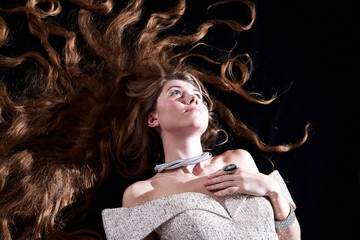 Obraz premium Young attractive girl with long blonde hair and beautiful dress posing lying on the floor in studio on dark background. Photo of the beautiful hair of model