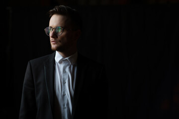 Portrait of handsome man dressed in suit and glasses looking away over black wall background.