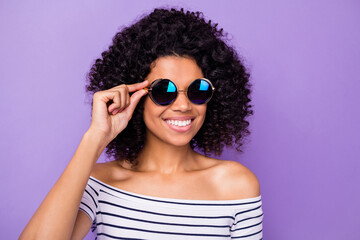 Portrait of charming cheerful dark skin girl hand touch sunglass beaming smile isolated on purple color background