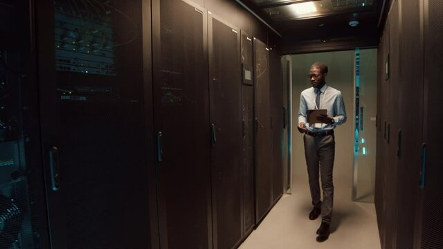 Wide shot of young African-American man wearing formalwear entering room with rack server cabinet with digital tablet in hands and starting analyzing work of computer