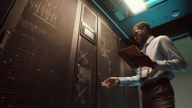 Low angle view of young African-American man wearing formalwear entering room with rack server cabinet with tablet in hands and starting checking work of computer