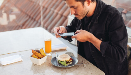 A young man taking picture with his smartphone of his meal - burger, roasted potato, beer on a terrace restaurant