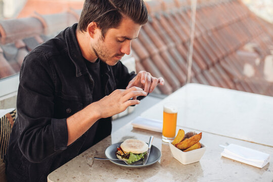 A young man taking picture with his smartphone of his meal - burger, roasted potato, beer on a terrace restaurant