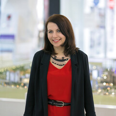 Portrait of a smiling woman in a red dress and black jacket in a light shopping center interior.