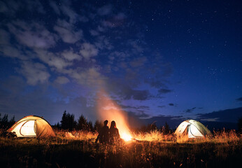 Fantastic view of night starry sky over grassy hill with illuminated camp tents, campfire and hikers. Young tourists having a rest near bonfire. Concept of hiking, night camping and relationship.