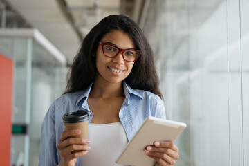 Beautiful smiling African American woman holding coffee cup and digital tablet looking at camera. Portrait of young confident businesswoman standing in office  