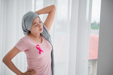 Fototapeta na wymiar Young asian women wearing hijab Open the curtains in the bedroom to get the morning sunshine. Attaching a pink ribbon represents recovery from a breast cancer patient. Breast cancer concept