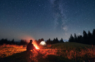 Silhouette of male traveler sitting near campfire under night sky with stars and Milky way. Starry sky under grassy hill with hiker, bonfire and camp tents. Concept of travelling and night camping. - obrazy, fototapety, plakaty