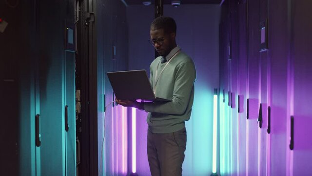 Medium shot of young African male IT administrator standing at rack server cabinet illuminated with neon light and using laptop to analyze its work