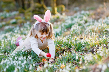 Little girl with Easter bunny ears making egg hunt in spring forest on sunny day, outdoors. Cute...